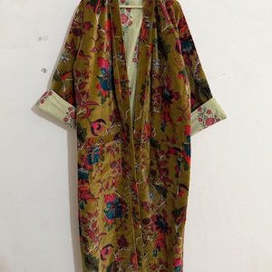 Op de afbeelding: Een lange, olijfgroene fluwelen kimono met een bloemenpatroon in rood, teal en roze. De kimono heeft een gestreepte voering en bloemenmanchetten. De kimono hangt aan een houten hanger.