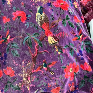 Peut inclure: Gros plan sur un tissu de velours violet vif avec un motif floral et d'oiseaux d&eacute;taill&eacute;. Le motif pr&eacute;sente des fleurs rouges et roses, des feuilles vertes et des oiseaux color&eacute;s perch&eacute;s sur des branches. Le tissu a un aspect doux et textur&eacute;.