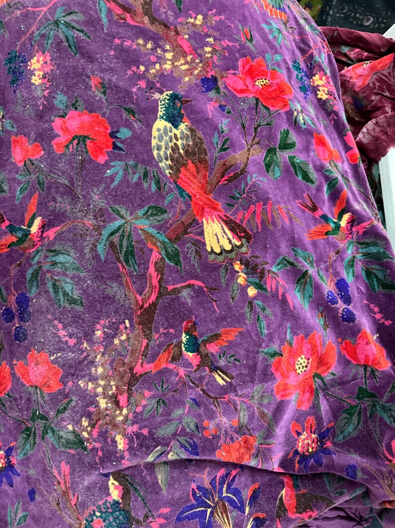 Peut inclure: Gros plan sur un tissu de velours violet vif avec un motif floral et d'oiseaux d&eacute;taill&eacute;. Le motif pr&eacute;sente des fleurs rouges et roses, des feuilles vertes et des oiseaux color&eacute;s perch&eacute;s sur des branches. Le tissu a un aspect doux et textur&eacute;.