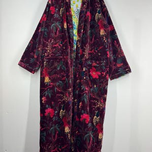 May include: A long, burgundy velvet robe with a floral and bird pattern. The robe has a contrasting patterned lining and two front pockets. The robe is hanging on a hanger.