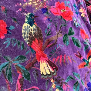 Peut inclure: Gros plan sur un tissu de velours violet avec un motif floral et d'oiseaux vibrant. Le motif pr&eacute;sente des oiseaux color&eacute;s perch&eacute;s sur des branches au milieu de grandes fleurs rouges et roses, de feuilles vertes et d'accents bleus. Le tissu a un aspect doux et textur&eacute;.