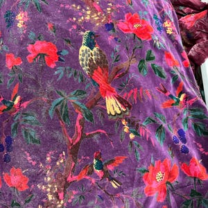 Peut inclure: Gros plan sur un tissu de velours violet avec un motif floral et d'oiseaux. Le motif pr&eacute;sente des fleurs rouges, des feuilles vertes et des oiseaux color&eacute;s perch&eacute;s sur des branches. Le tissu a un aspect doux et textur&eacute;.