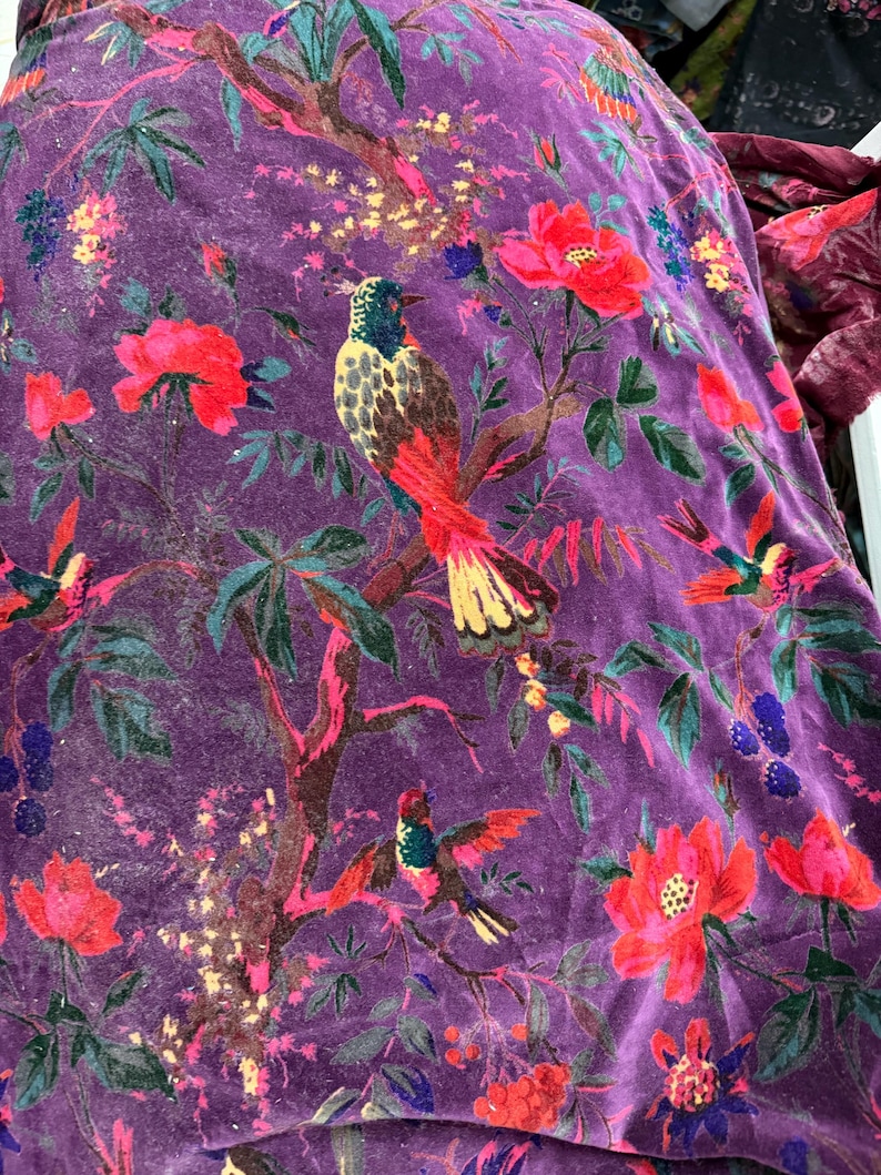 Peut inclure: Gros plan sur un tissu de velours violet avec un motif floral et d'oiseaux. Le motif pr&eacute;sente des fleurs rouges, des feuilles vertes et des oiseaux color&eacute;s perch&eacute;s sur des branches. Le tissu a un aspect doux et textur&eacute;.