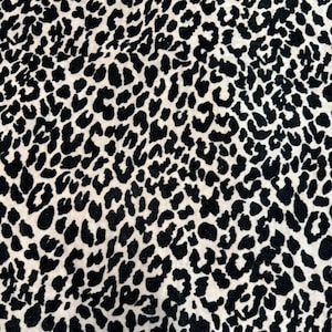 Puede incluir: Primer plano de una tela con un estampado de leopardo en negro y crema. El patrón presenta manchas negras irregulares de varios tamaños dispersas sobre un fondo crema. La tela parece suave y texturizada.