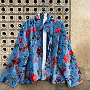 Könnte beinhalten: Hellblaue Samtjacke mit floralem Muster mit roten Blumen und Vögeln. Die Jacke hat ein gestreiftes Futter und hängt an einem Metallbügel.