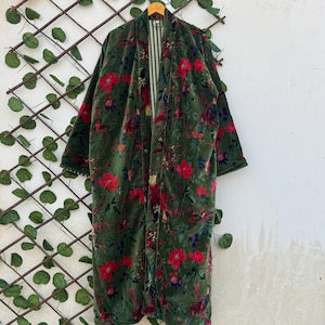 Może przedstawiać: Długa, rozpinana szata z ciemnozielonego aksamitu, ozdobiona żywym kwiatowym wzorem z czerwonymi i niebieskimi kwiatami. Szata wisi na drewnianym wieszaku na białej ścianie z dekoracyjną kratą.