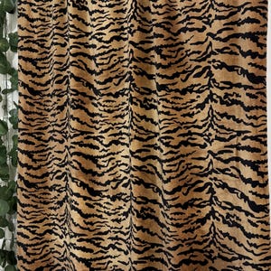May include: A curtain with a tan and black tiger stripe pattern. The fabric is draped, displaying vertical lines of the tiger stripe design. The curtain hangs from a rod, with a hint of greenery on the left.