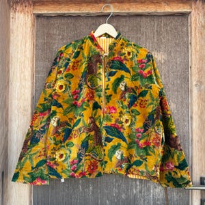 Może przedstawiać: Żółta aksamitna kurtka z żywym nadrukiem kwiatowym i małpim. Kurtka posiada zamek błyskawiczny z przodu, długie rękawy i kołnierz mandaryński. Podszewka ma żółto-biały wzór w paski.