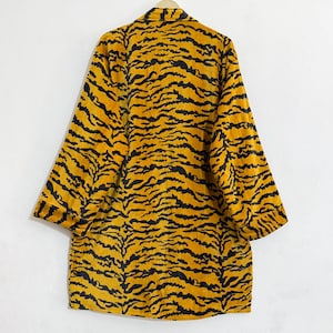 May include: A long-sleeved jacket featuring a bold mustard yellow and black tiger stripe pattern. The garment has a classic collar, a button-down front, and wide sleeves with cuffs, displayed hanging on a hanger against a plain white wall.