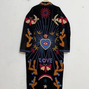 May include: A black long-sleeve coat featuring colourful embroidery of a central heart with an eye, the word LOVE, moths, cacti, a sun, and a rainbow. The design includes blue radiating lines and small stars, with a collar detail and long sleeves.