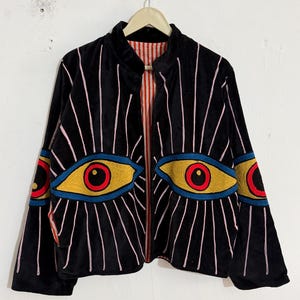 May include: A black jacket featuring an embroidered eye design on the front and sleeves. The eyes have yellow irises, red pupils, and blue outlines, with pink embroidered lines radiating outward. The jacket has a stand collar and a red and white striped lining.