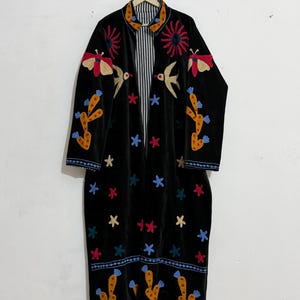 May include: A black long-sleeve coat featuring colorful embroidered motifs including yellow cacti with blue flowers, red suns, moths, birds, and scattered stars. The garment has a collar and a black and white vertical striped lining visible inside.