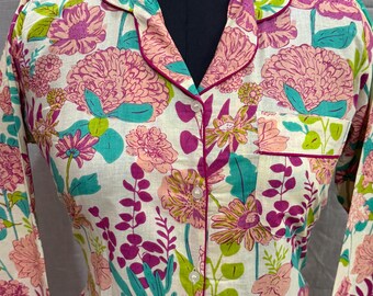 Könnte beinhalten: Ein floraler Pyjama-Set mit cremefarbenem Hintergrund, verziert mit rosa, lila und türkisfarbenen Blumen und grünen Blättern. Das langärmlige Oberteil hat einen Reverskragen und eine Tasche, alles mit einem rosa Rand versehen.