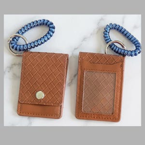 May include: Two brown leather cardholders with a woven pattern, each attached to a blue braided keychain. One cardholder has a snap closure, while the other has a clear window to display an ID or card.