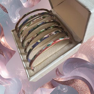 May include: A set of colorful enamel bangles in a box. The bangles feature various designs with red, blue, green, and brown accents. The box is open, revealing the collection of bracelets. The background has a soft, abstract design with shades of pink and purple.
