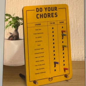 May include: A yellow rectangular chore chart with the words "DO YOUR CHORES" at the top. The chart lists chores such as "Make bed" and "Clean bathroom." It has a "TO DO" and "DONE" section with colorful pegs to mark progress. A small potted plant is in the background.