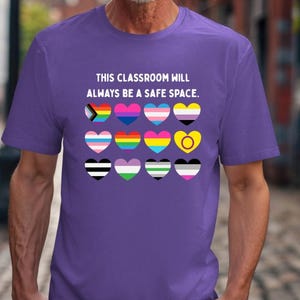 Puede incluir: Camiseta morada con el texto "This classroom will always be a safe space." y una fila de corazones con diferentes banderas del orgullo dentro.
