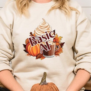 Peut inclure: Sweat-shirt beige avec un motif de latte à la citrouille épicée, des citrouilles et des feuilles d'automne. Le texte "Basic AF" est imprimé sur le devant. Une citrouille orange décorative est tenue sur les genoux.