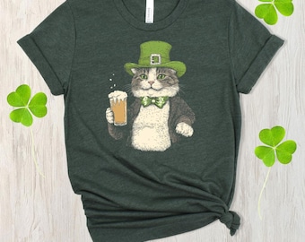 May include: Dark green t-shirt with a cartoon cat wearing a green hat and bow tie, holding a pint of beer. The cat is dressed in a suit jacket. The shirt has a rolled sleeve and a knot at the bottom. Shamrock accents are in the background.