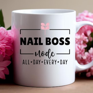 Könnte beinhalten: Weiße Keramiktasse mit schwarzem Text, der "NAIL BOSS mode ALL DAY EVERY DAY" lautet. Eine kleine rosa Blumengrafik befindet sich über dem Text.