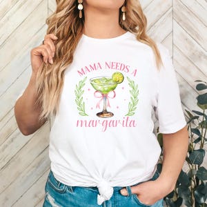 Op de afbeelding: Wit T-shirt met de tekst "Mama Needs a Margarita" in roze letters. In het midden van de tekst is een groen margarita-glas met een limoenpartje afgebeeld.