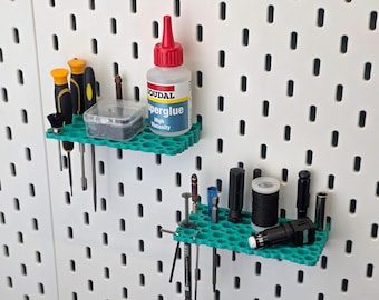 May include: Two green, honeycomb-patterned shelves display tools and supplies against a white pegboard. A bottle of superglue with a red cap is on the top shelf. Screwdrivers, bits, and other small tools are also visible.