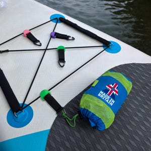Pode incluir: Uma prancha de paddleboard branca e azul com um sistema de cordas elásticas pretas e uma bolsa azul e verde. A bolsa tem o texto "MADE IN BRITAIN" e uma bandeira do Reino Unido.