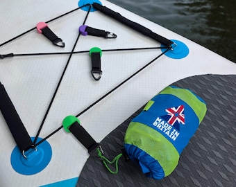 Puede incluir: Tabla de paddle surf blanca y azul con un sistema de cuerdas elásticas negras y una bolsa azul y verde. La bolsa tiene el texto "MADE IN BRITAIN" y una bandera del Reino Unido.