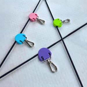 Pode incluir: Quatro fechos de cordão redondos e coloridos em rosa, azul, verde e roxo, cada um com um fecho de metal prateado. Os fechos de cordão estão presos a cordões pretos, dispostos em um padrão geométrico.
