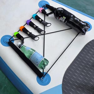 Pode incluir: Uma prancha de paddleboard branca e azul com um sistema de corda elástica preta e suportes de garrafa coloridos. Uma garrafa preta e uma garrafa verde são fixadas pelos suportes. Os suportes são em verde, laranja, roxo, azul e rosa.