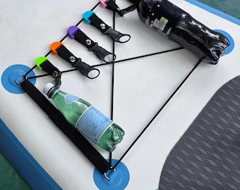 Könnte beinhalten: Ein weißes und blaues Paddleboard mit einem schwarzen Bungee-Kordel-System und farbigen Flaschenhaltern. Eine schwarze Flasche und eine grüne Flasche werden von den Haltern gehalten. Die Halter sind in Grün, Orange, Lila, Blau und Rosa.