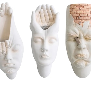 May include: Three white ceramic wall planters shaped like faces. The first planter has a hand covering the face, the second planter has a hand covering the top of the head, and the third planter has a cracked brick wall covering the top of the head.