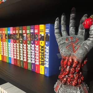May include: A complete set of 16 Berserk manga volumes, with the spines featuring the title "Berserk" and the author's name "Kentaro Miura". A hand sculpture with a red ornament is in the foreground.
