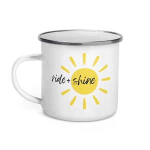 以下が含まれることがあります： 白いエナメルマグに、黄色の太陽と黒い文字で「ride + shine」と書かれています。