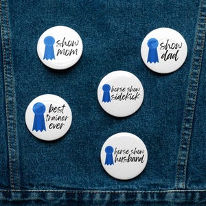 May include: A collection of white circular badges featuring blue ribbon designs and text. The badges read "Show Mom", "Show Dad", "Best Trainer Ever", "Horse Show Sidekick", and "Horse Show Husband". Set against a blue denim backdrop.