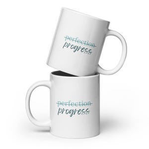 May include: Two white ceramic mugs stacked on top of each other. Both mugs feature the text "perfection progress" with a line through the word "perfection". The text is in a blue and green gradient.