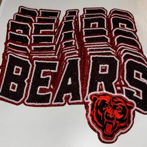May include: A collection of stacked fabric patches featuring the word BEARS in black block letters with a white sequin border. One patch at the bottom right includes a detailed embroidered graphic of a snarling bear head in red and black.
