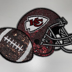 May include: A decorative patch featuring a football and a football helmet covered in red, brown, white, and silver sequins. The helmet displays a white arrowhead logo with the red letters KC. The design is outlined in silver glitter trim.