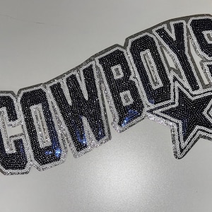 May include: A decorative patch featuring the word COWBOYS in dark blue sequins with a silver sequin outline, accompanied by a five-pointed star symbol also rendered in dark blue and silver sequins, designed for application to fabric surfaces.