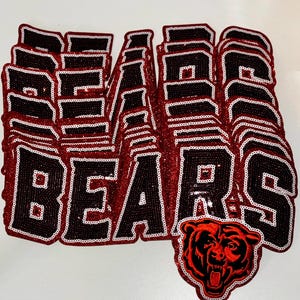 May include: A stack of multiple iron-on patches featuring the word BEARS in black block letters with a white outline and a red glitter border. The bottom patch includes a detailed graphic of a roaring bear head in red and black glitter.