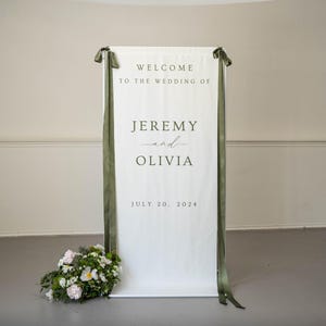 K&ouml;nnte beinhalten: Ein wei&szlig;es Hochzeits-Willkommensschild mit dem Text "WELCOME TO THE WEDDING OF JEREMY and OLIVIA JULY 20, 2024". Das Schild ist mit olivgr&uuml;nen B&auml;ndern und einem Blumenarrangement verziert.
