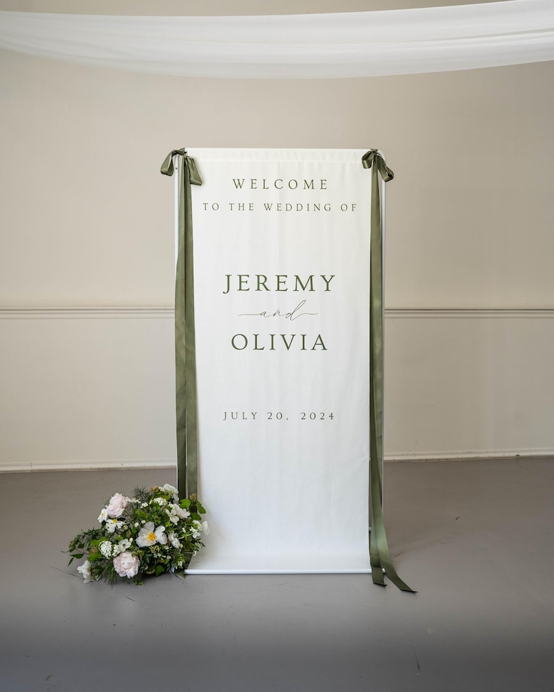 K&ouml;nnte beinhalten: Ein wei&szlig;es Hochzeits-Willkommensschild mit dem Text "WELCOME TO THE WEDDING OF JEREMY and OLIVIA JULY 20, 2024". Das Schild ist mit olivgr&uuml;nen B&auml;ndern und einem Blumenarrangement verziert.