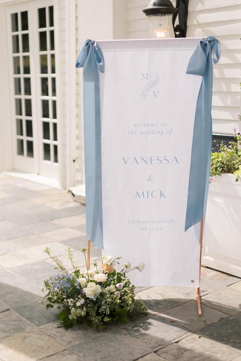 K&ouml;nnte beinhalten: Ein wei&szlig;es Hochzeits-Willkommensschild mit blauen B&auml;ndern und kupferfarbenen St&uuml;tzen. Auf dem Schild steht "welcome to the wedding of VANESSA & MICK" mit Ort und Datum. Am Fu&szlig; befindet sich ein Blumengesteck.
