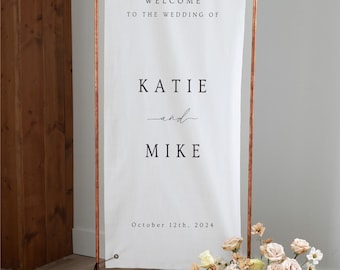 May include: A tall white fabric wedding welcome sign suspended on a copper pipe frame. The text reads: WELCOME TO THE WEDDING OF KATIE and MIKE October 12th, 2024. A floral arrangement with light pink, cream, and white roses sits at the base.