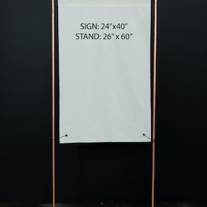 K&ouml;nnte beinhalten: Ein kupferfarbener Metall-Schilderst&auml;nder mit einem wei&szlig;en Schild. Das Schild ist 61 cm x 102 cm gro&szlig; und der St&auml;nder ist 66 cm x 152 cm gro&szlig;. Das Schild ist mit schwarzem Faden am St&auml;nder befestigt. Der Hintergrund ist schwarz.