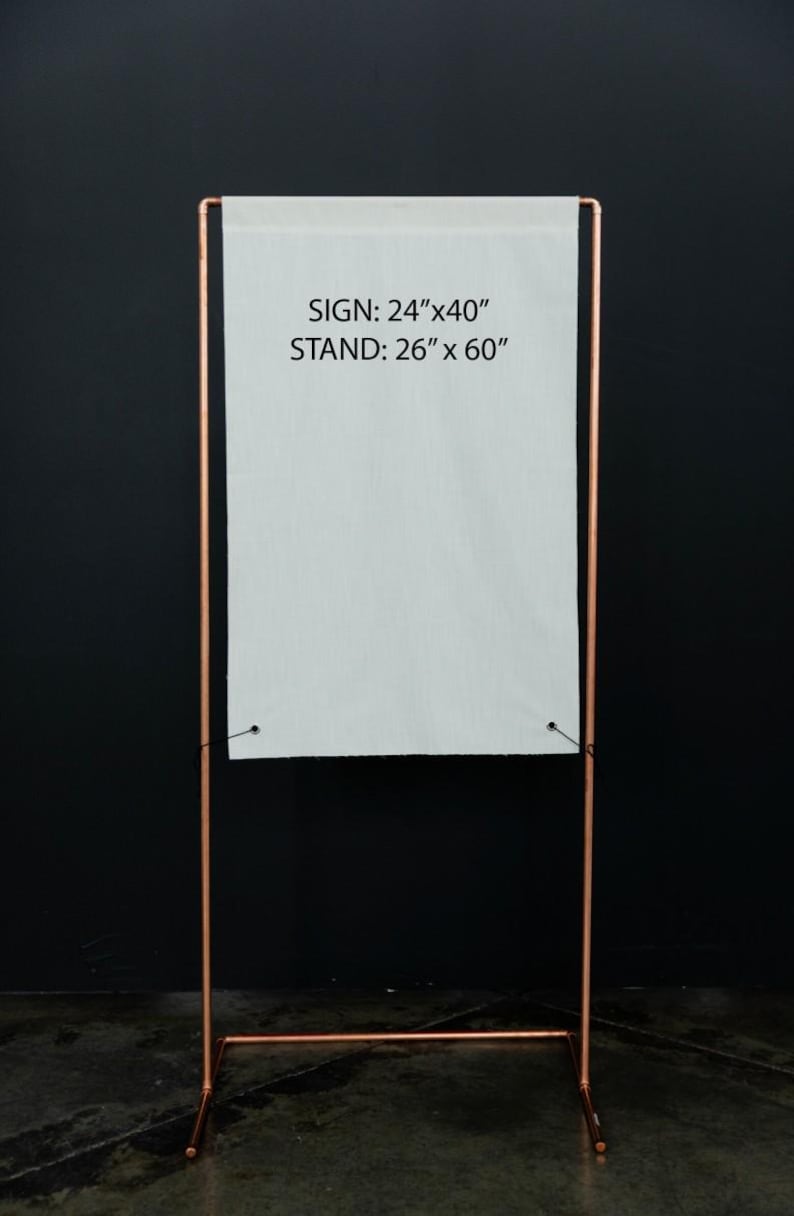 K&ouml;nnte beinhalten: Ein kupferfarbener Metall-Schilderst&auml;nder mit einem wei&szlig;en Schild. Das Schild ist 61 cm x 102 cm gro&szlig; und der St&auml;nder ist 66 cm x 152 cm gro&szlig;. Das Schild ist mit schwarzem Faden am St&auml;nder befestigt. Der Hintergrund ist schwarz.