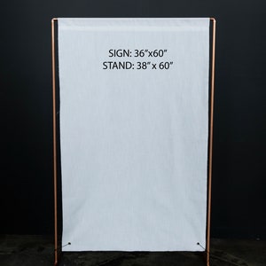 K&ouml;nnte beinhalten: Ein wei&szlig;es Stoffschild, 91 cm x 152 cm, wird auf einem kupferfarbenen Metallst&auml;nder, 96 cm x 152 cm, pr&auml;sentiert. Das Schild und der St&auml;nder befinden sich vor einem dunklen Hintergrund. Der Text "SIGN: 36"x60" STAND: 38" x 60"" ist sichtbar.