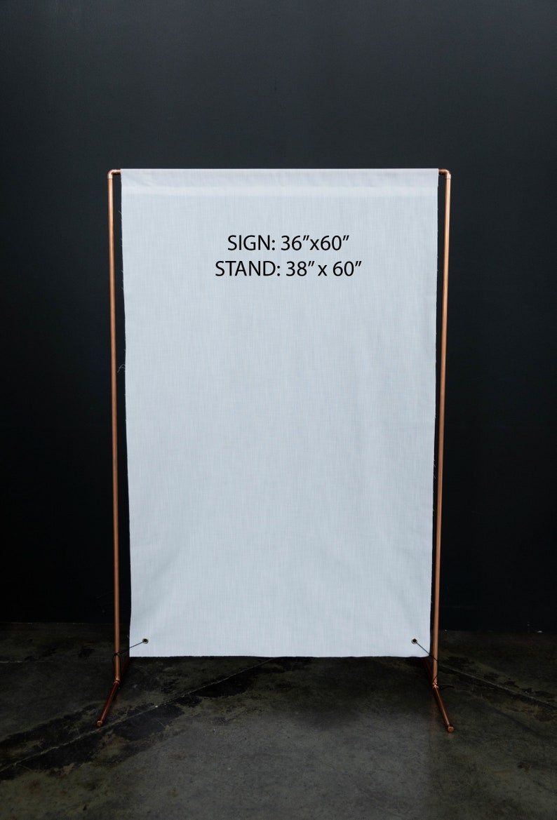 K&ouml;nnte beinhalten: Ein wei&szlig;es Stoffschild, 91 cm x 152 cm, wird auf einem kupferfarbenen Metallst&auml;nder, 96 cm x 152 cm, pr&auml;sentiert. Das Schild und der St&auml;nder befinden sich vor einem dunklen Hintergrund. Der Text "SIGN: 36"x60" STAND: 38" x 60"" ist sichtbar.