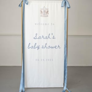 May include: A white vertical banner with blue ribbon ties hanging from a gold metal frame. The banner features a carousel illustration at the top and the text "welcome to Sarah's baby shower" followed by the date "06.28.2025" in blue lettering.