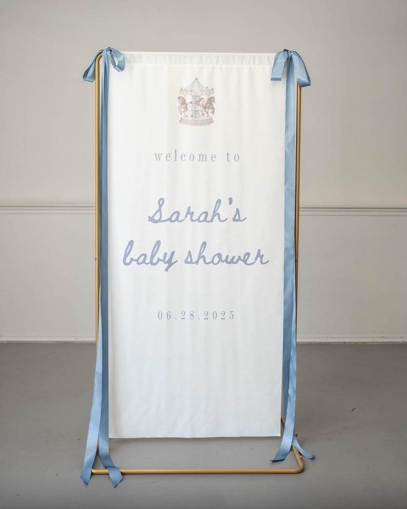 May include: A white vertical banner with blue ribbon ties hanging from a gold metal frame. The banner features a carousel illustration at the top and the text "welcome to Sarah's baby shower" followed by the date "06.28.2025" in blue lettering.